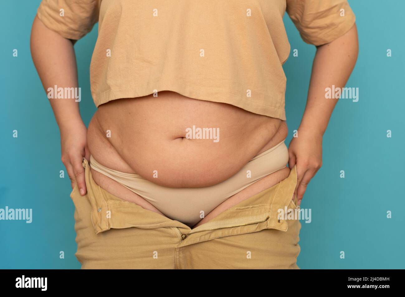 Unrecognizable plump woman wearing beige T-shirt, pulling on unzipped jeans, showing excess belly on blue background. Stock Photo