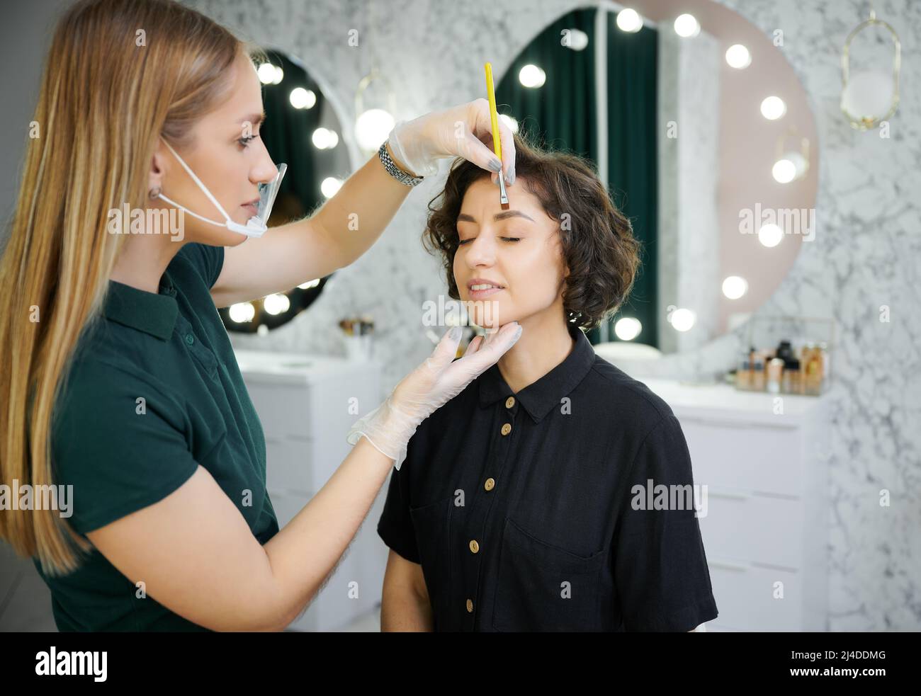 Female beauty specialist shaping client eyebrows with cosmetic brow brush. Young woman keeping eyes closed and smiling while having eyebrow correction procedure in beauty salon. Stock Photo