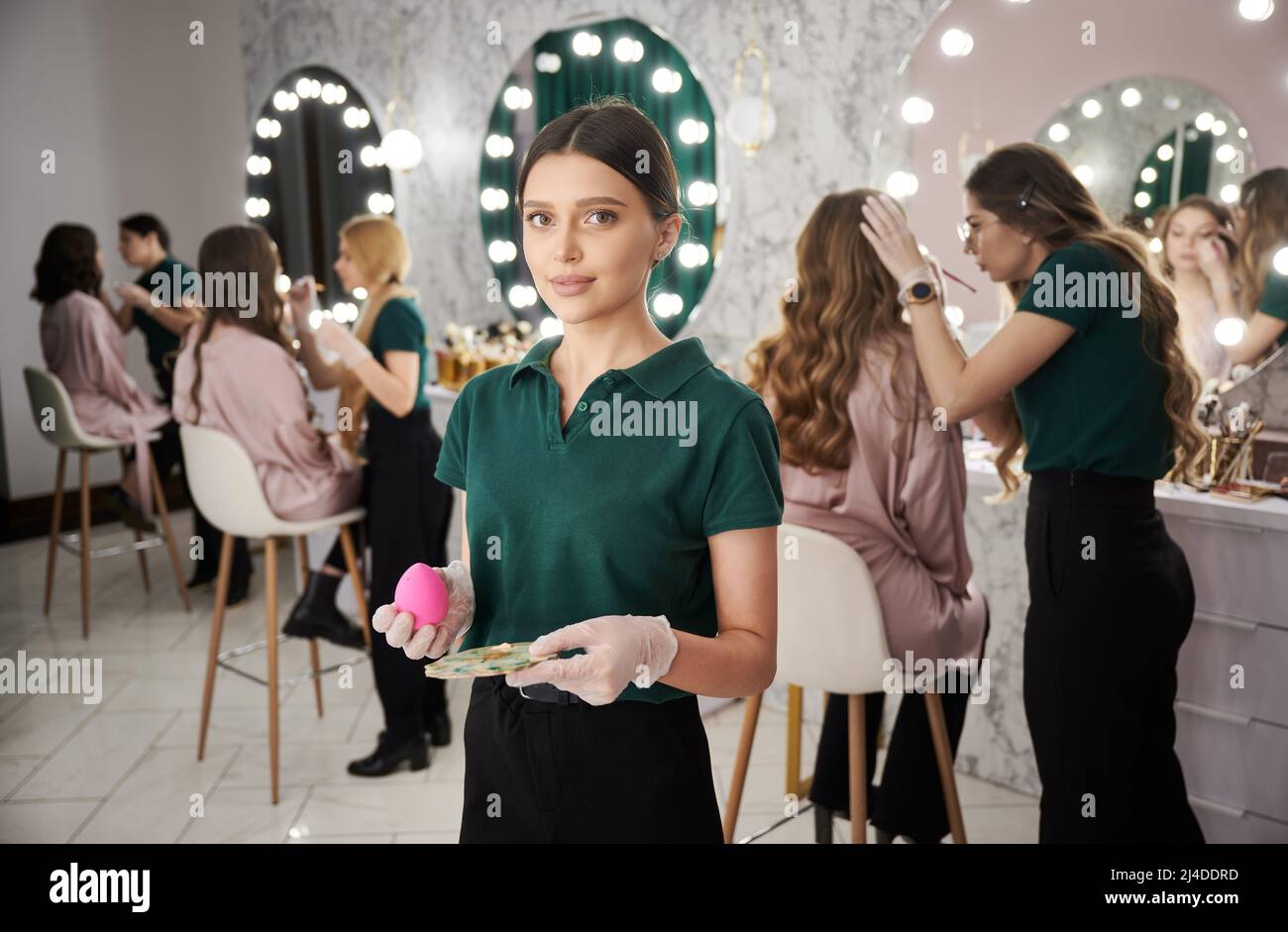 Portrait of beautiful young woman in gloves looking at camera and smiling while holding cosmetic sponge. Makeup artist standing in beauty salon while colleagues doing makeup for female clients. Stock Photo