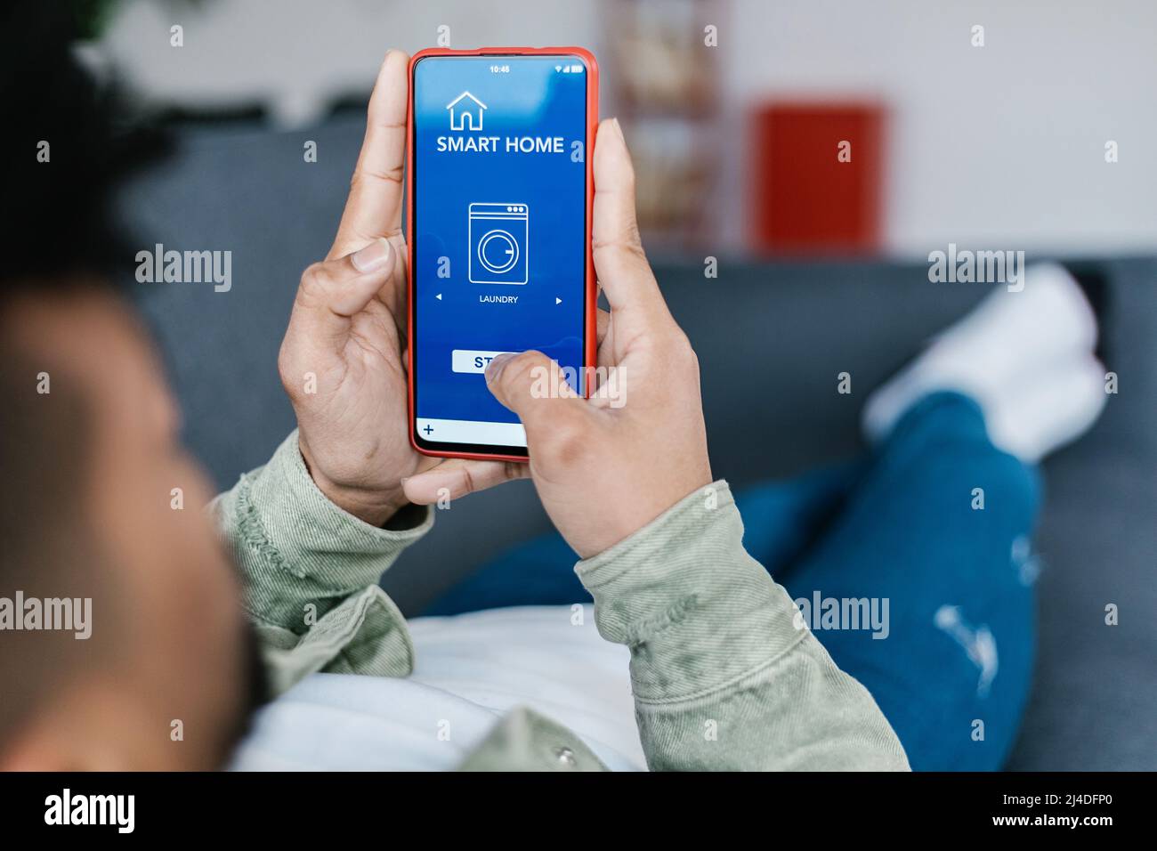 Young man lying on sofa and holding phone with app for controlling smart home Stock Photo