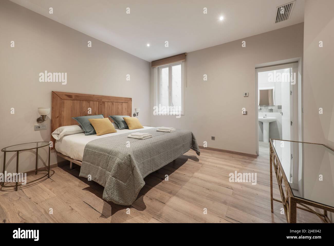 Bedroom with a large bed with white sheets, a wooden headboard, green and yellow cushions, an en-suite toilet and chestnut parquet flooring Stock Photo