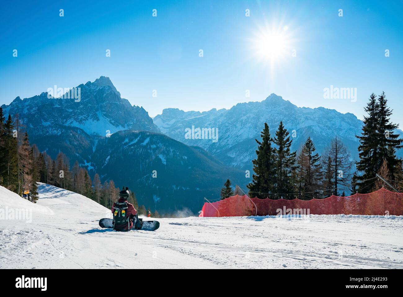 Sun shining on tired snowboarder sitting on snowy landscape against mountains Stock Photo