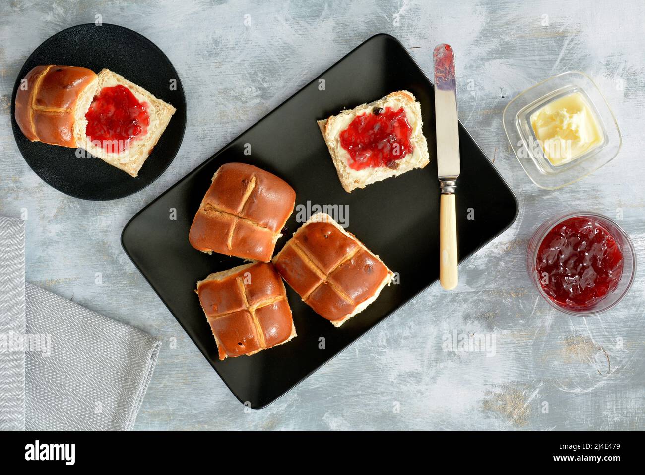 Fresh baked hot cross buns with raspberry jam and butter on black matte plates shot from overhead.  Traditional Easter baked treat. Stock Photo