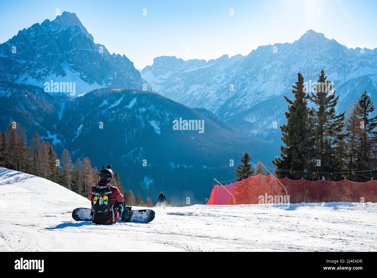 Rear view of tired snowboarder sitting on snowy landscape against mountains Stock Photo