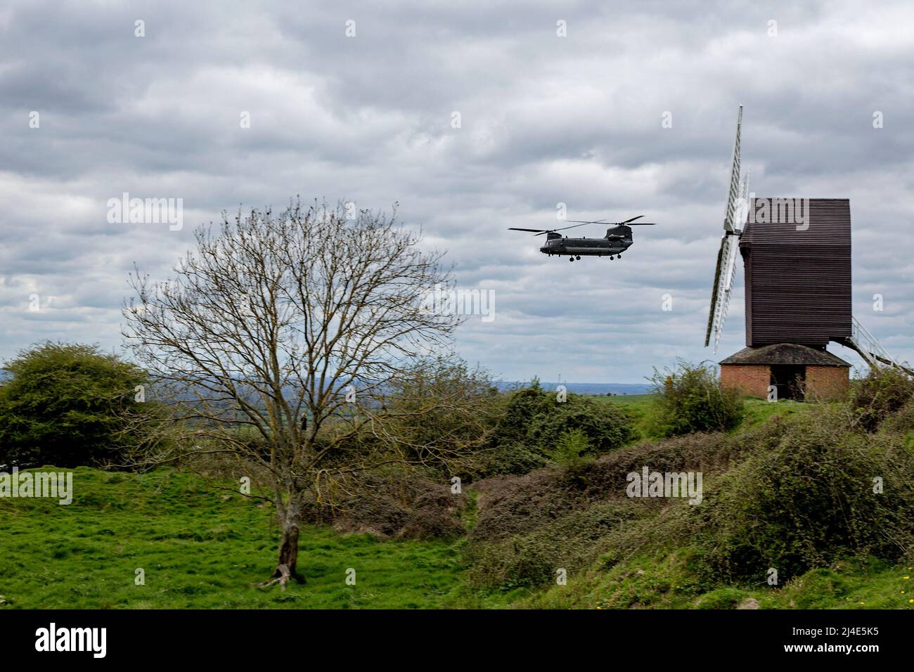 A Royal Air Force CH-47 Chinook helicopter flies low and close past the windmill at Brill, Buckinghamshire, UK on a spring day with low grey clouds. Stock Photo