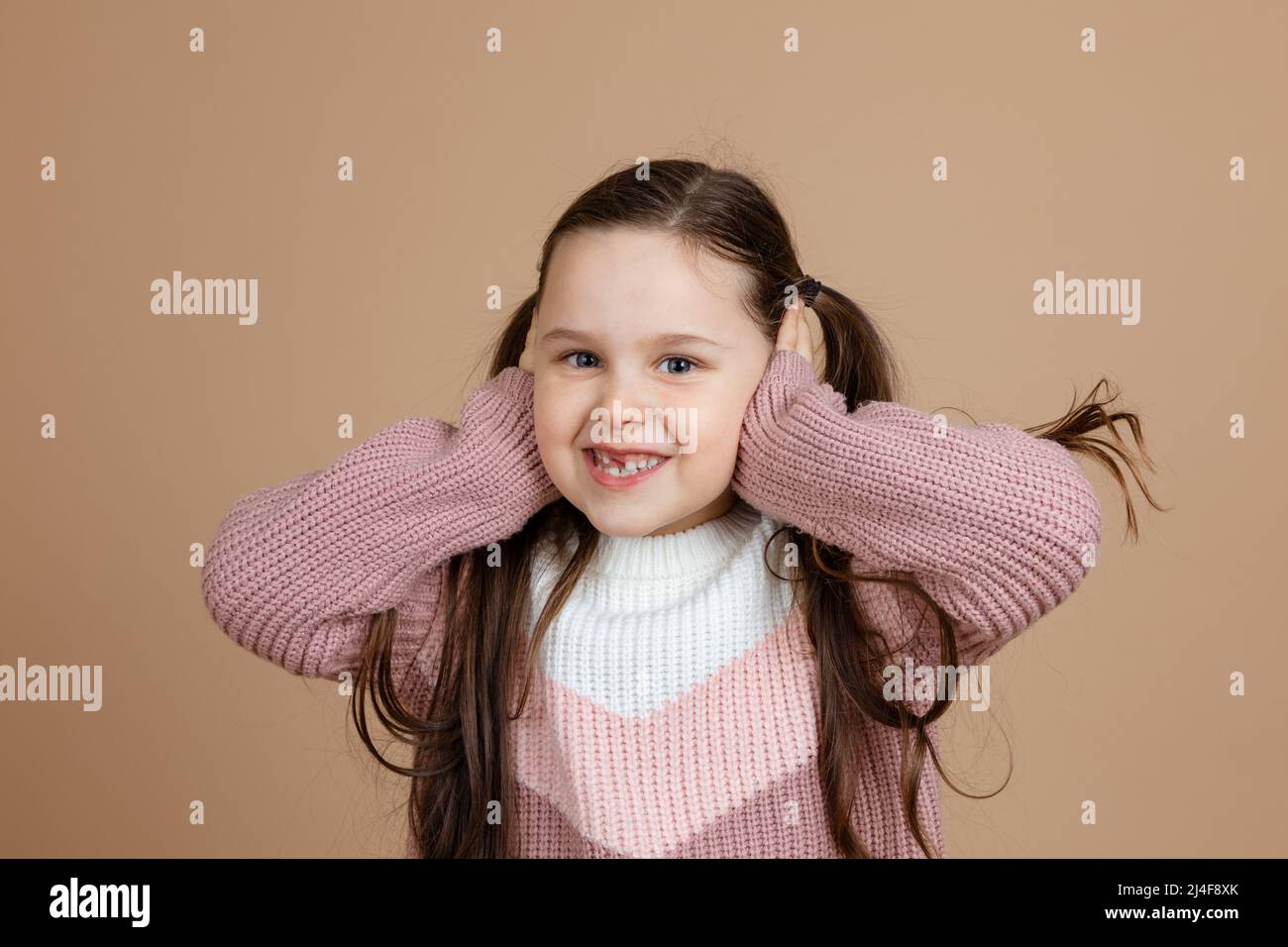 Portrait of young awesome pretty smiling girl with long dark hair in white, pink sweater standing, covering ears with hands, posing.  Stock Photo