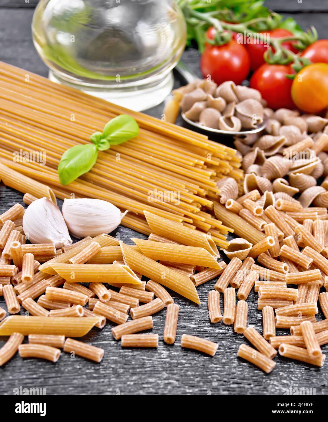 Pasta whole grain and rye pipe rigate, spaghetti, penne; seashells, rigatoni, tomatoes on a branch, garlic, vegetable oil in a decanter on black woode Stock Photo