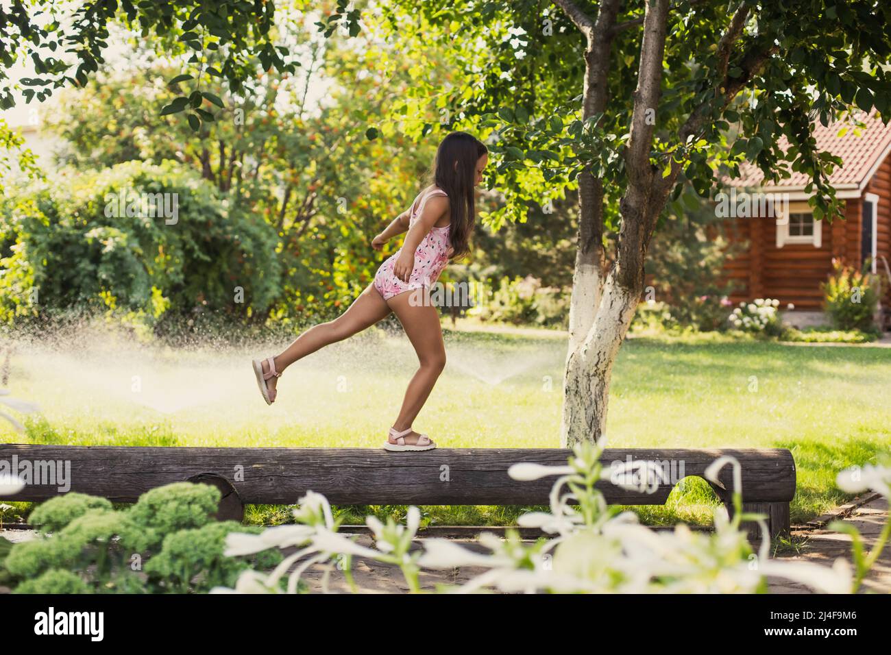 Female kid balancing on log on one foot looking like swallow indulging with water sprinklers, house and green trees in background in daytime. Dreaming Stock Photo