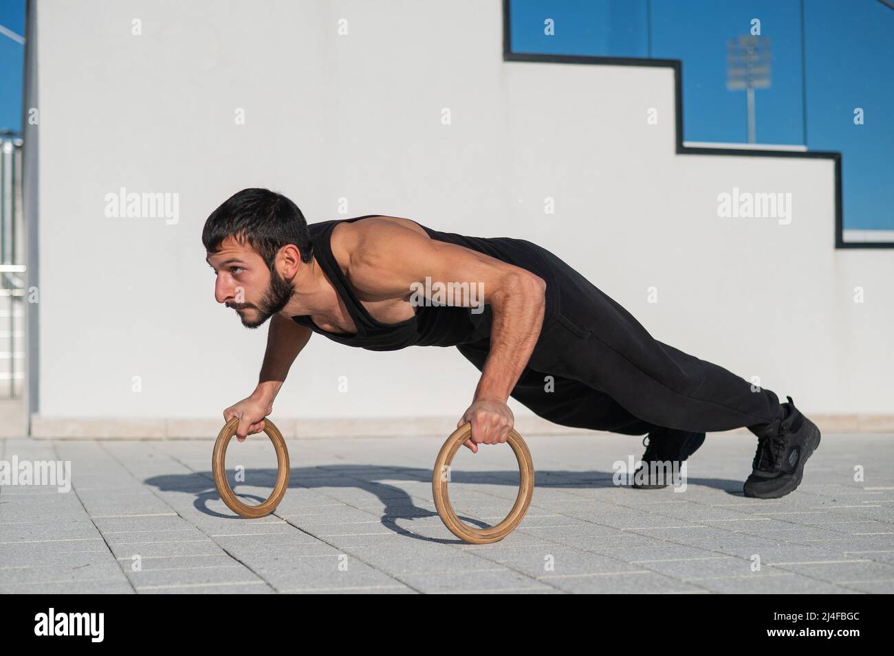 A man in black sportswear doing ring push-ups outdoors. Stock Photo