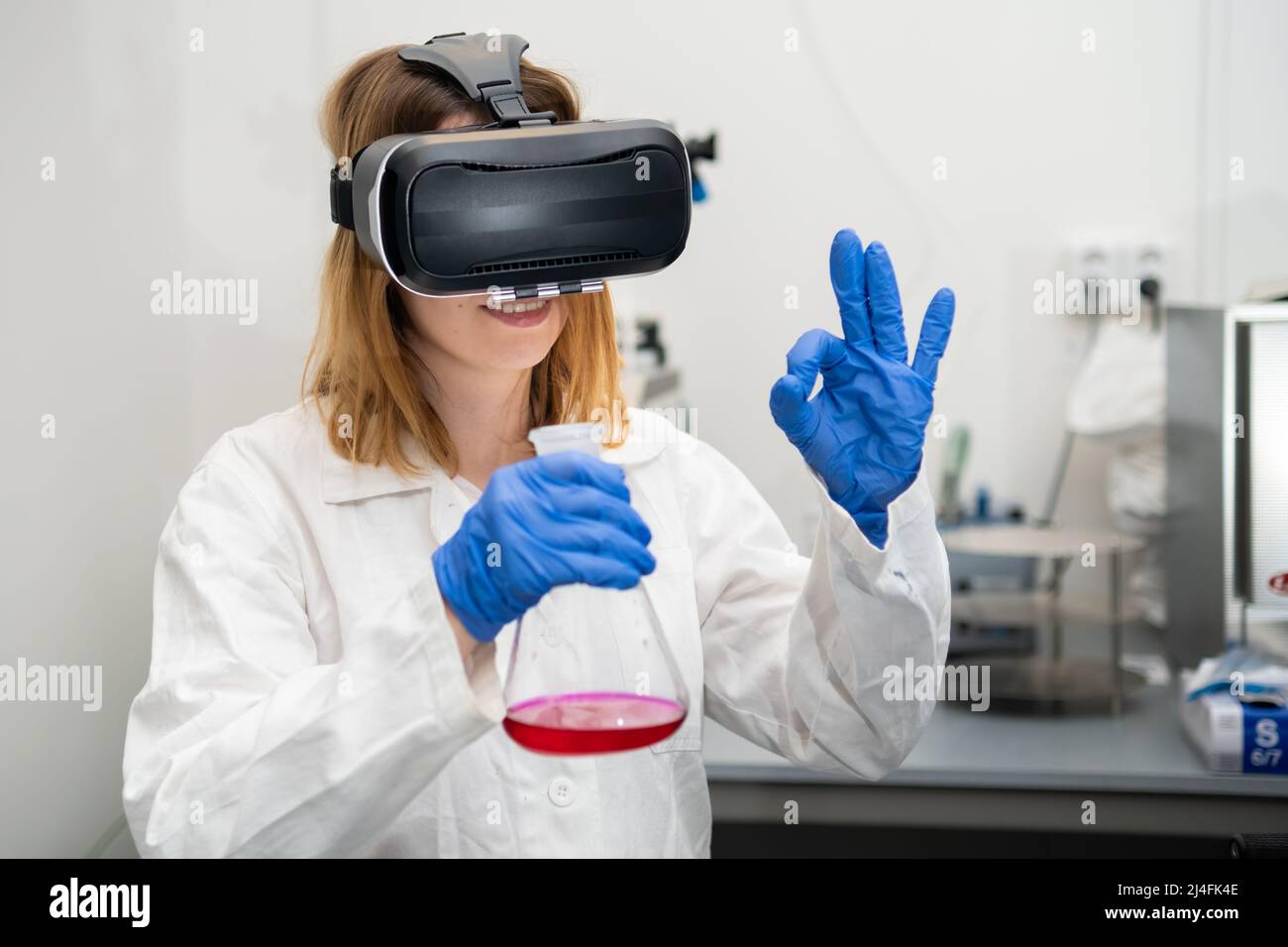 Female scientist show sign OK in VR glass, playing metaverse NFT game. Metaverse virtual world for future science and research.  Stock Photo