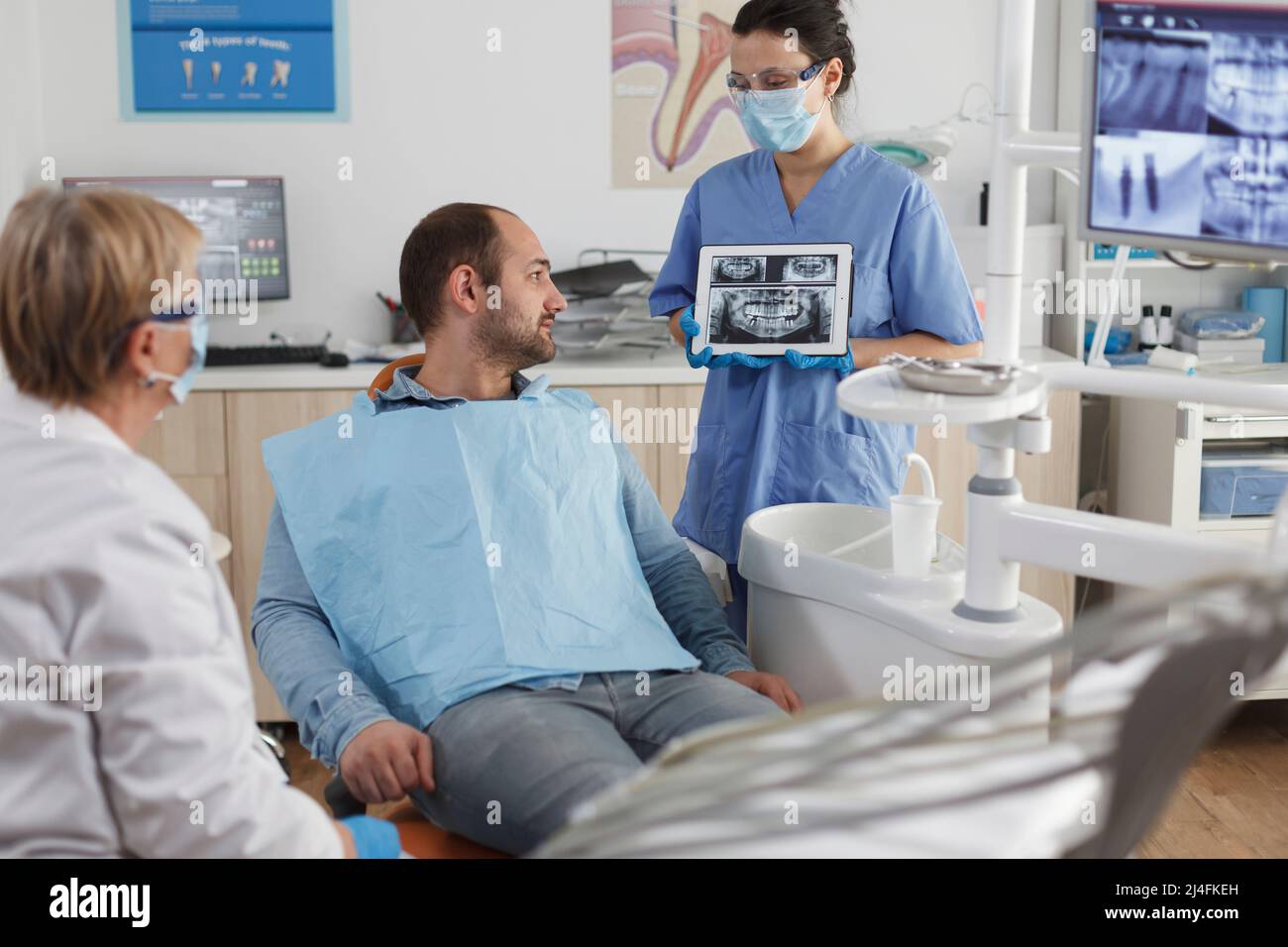 Dentist assistant with face mask holding tabet computer explaining teeth radiography to sick