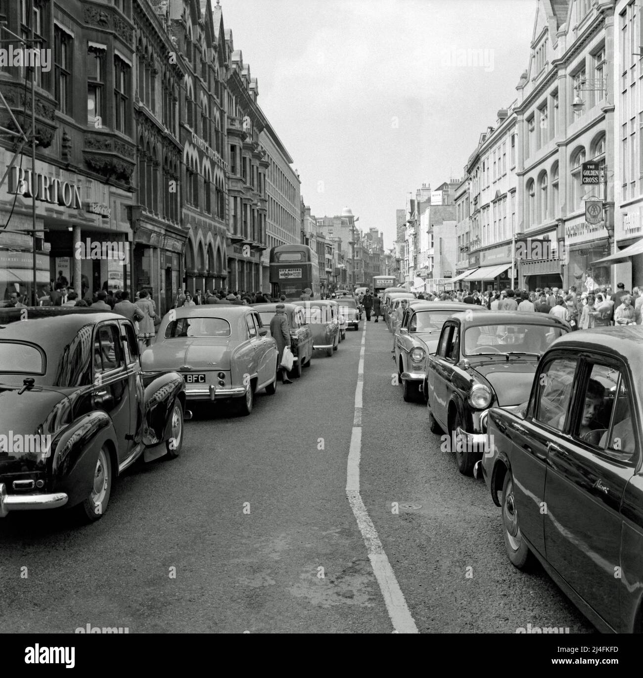 A very busy city centre view in Oxford – a view north looking up Cornmarket Street, Oxford, Oxfordshire, England, UK from near its junction with the High Street in 1960. At that time the street was very car-orientated and their appears to be a traffic jam with cars at a standstill. A policeman is in the centre of the road. Shoppers are in abundance too. Today the street has been largely pedestrianised – a vintage 1960s photograph. Stock Photo