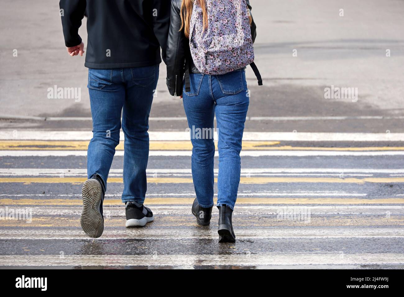 Couple in jeans walks on pedestrian crossing holding hands. Road zebra marking, people on wet crosswalk in spring weather, street safety concept Stock Photo