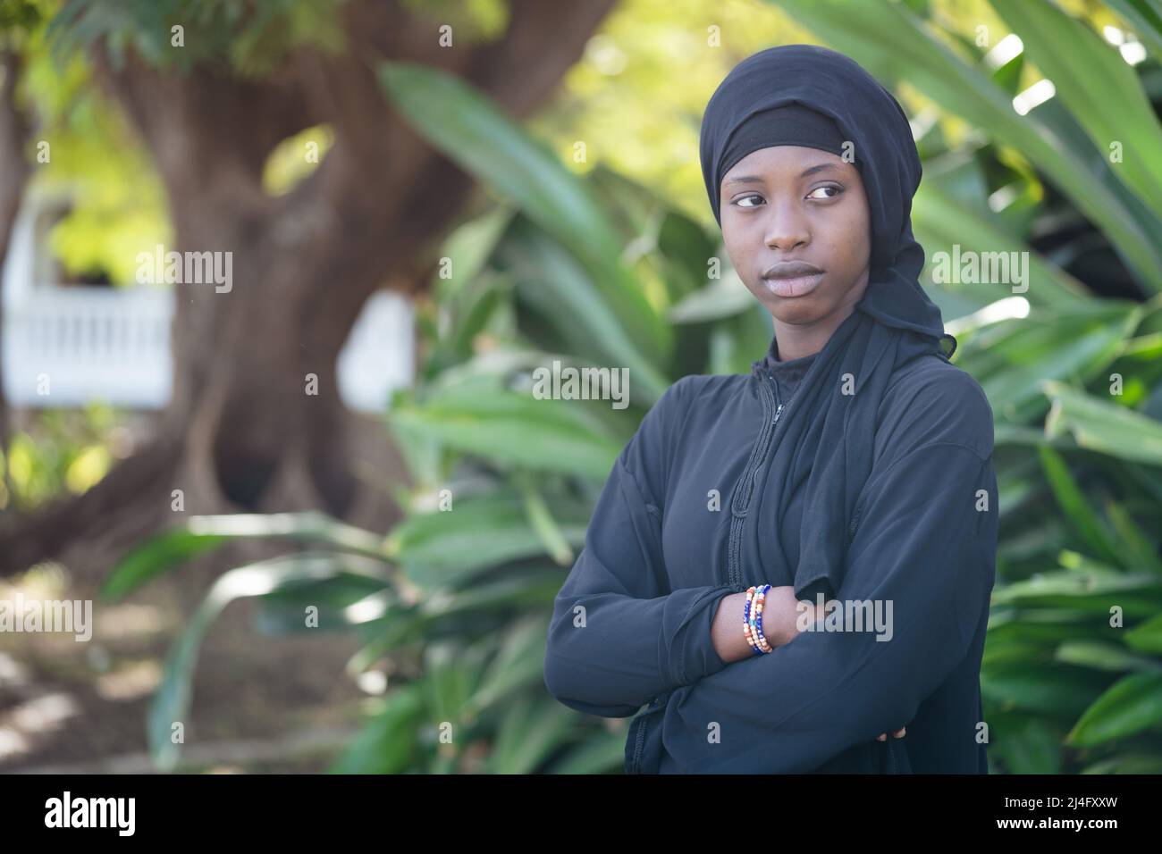 Outdoor shot of a pensive young African girl with her arms crossed in strict black clothing with a headscarf, symbolising troubles and doubts of adole Stock Photo
