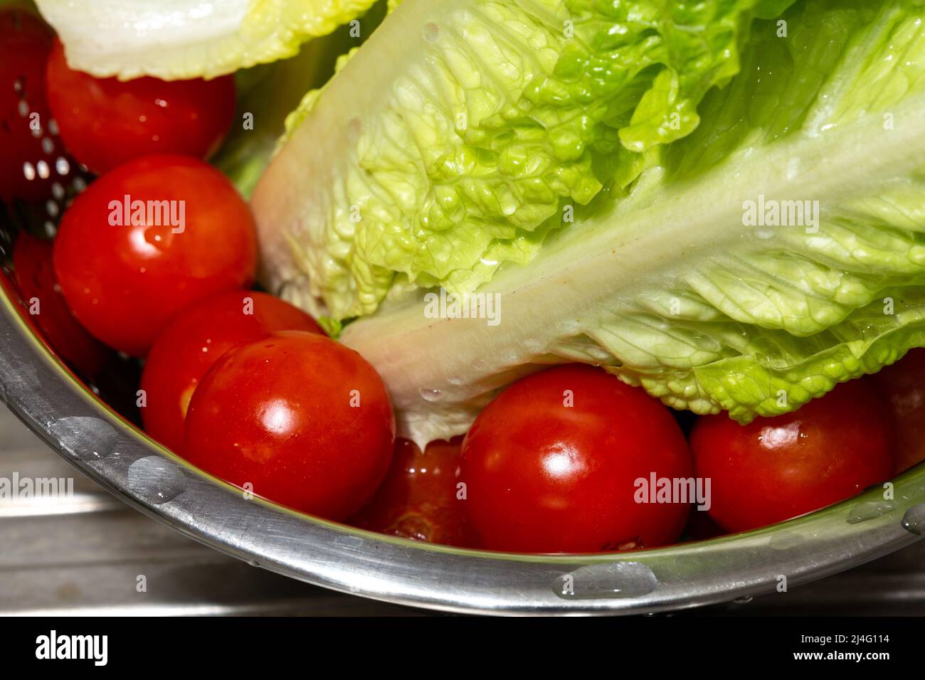 Close-up of small fresh tomatoes and lettuce leaves washed in a metal strainer. Tomatoes on the bottom in selective focus. Stock Photo