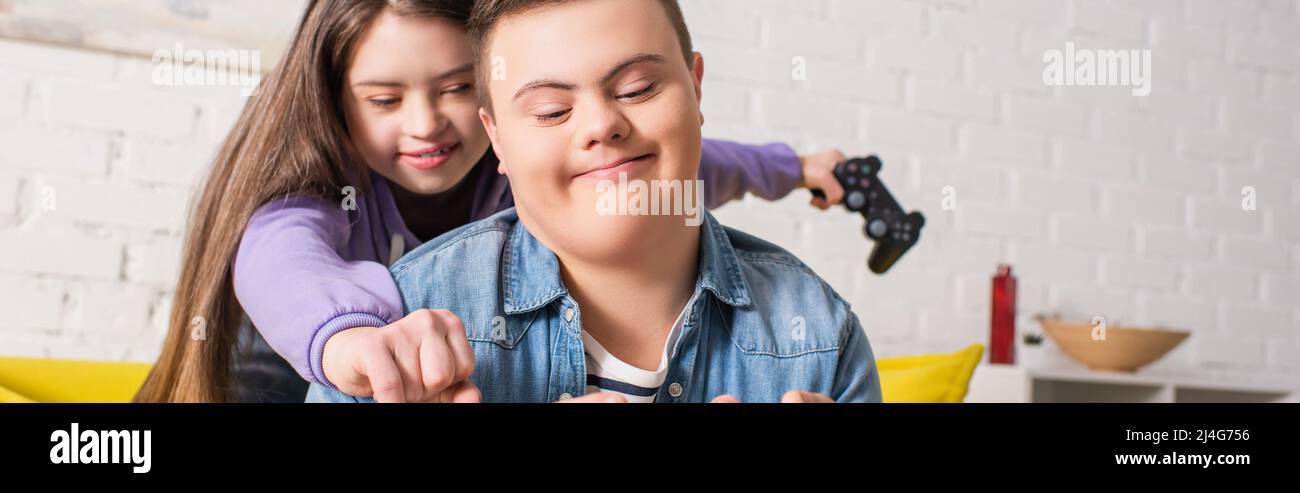 KYIV, UKRAINE - JANUARY 21, 2022: Teen girl with down syndrome holding blurred joystick near friend at home, banner Stock Photo