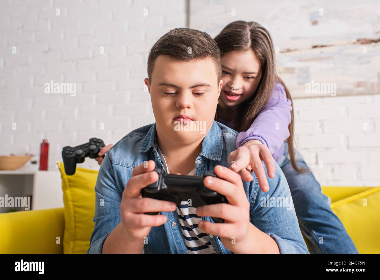 KYIV, UKRAINE - JANUARY 21, 2022: Girl with down syndrome pointing at joystick in hand of teen friend at home Stock Photo