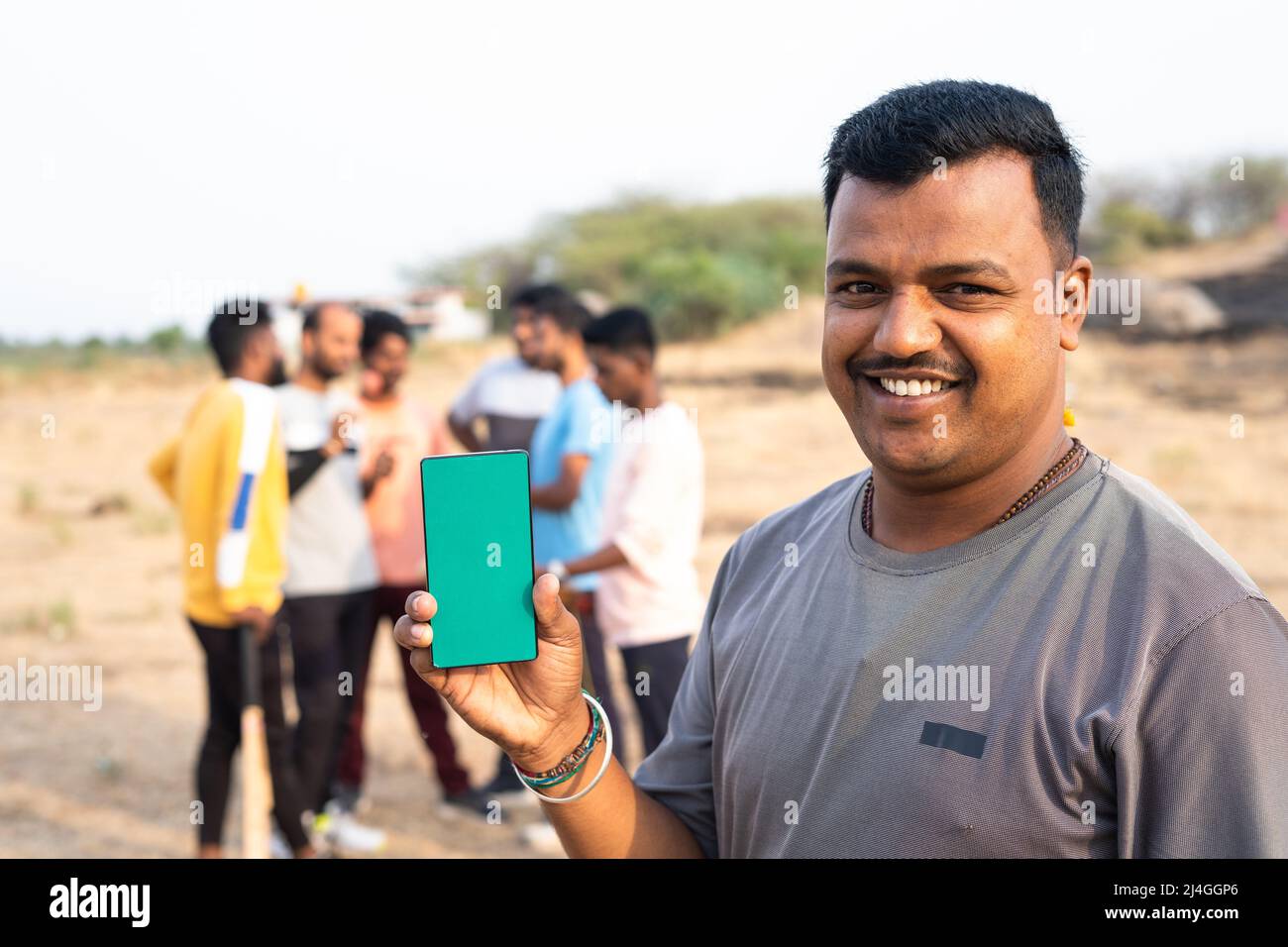 Smiling cricket player showing mobile phone by looking camera at playground in front of players - concept of online booking, app promotion and Stock Photo