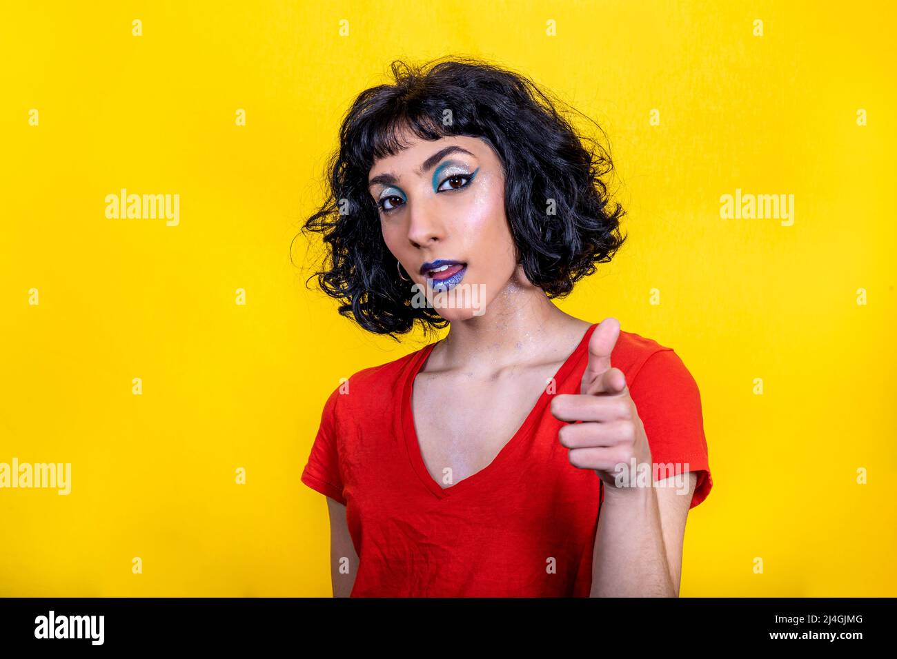Smiling young woman in red t-shirt is pointing at you. Woman portrait with trendy look and bright colors on yellow background. Stock Photo