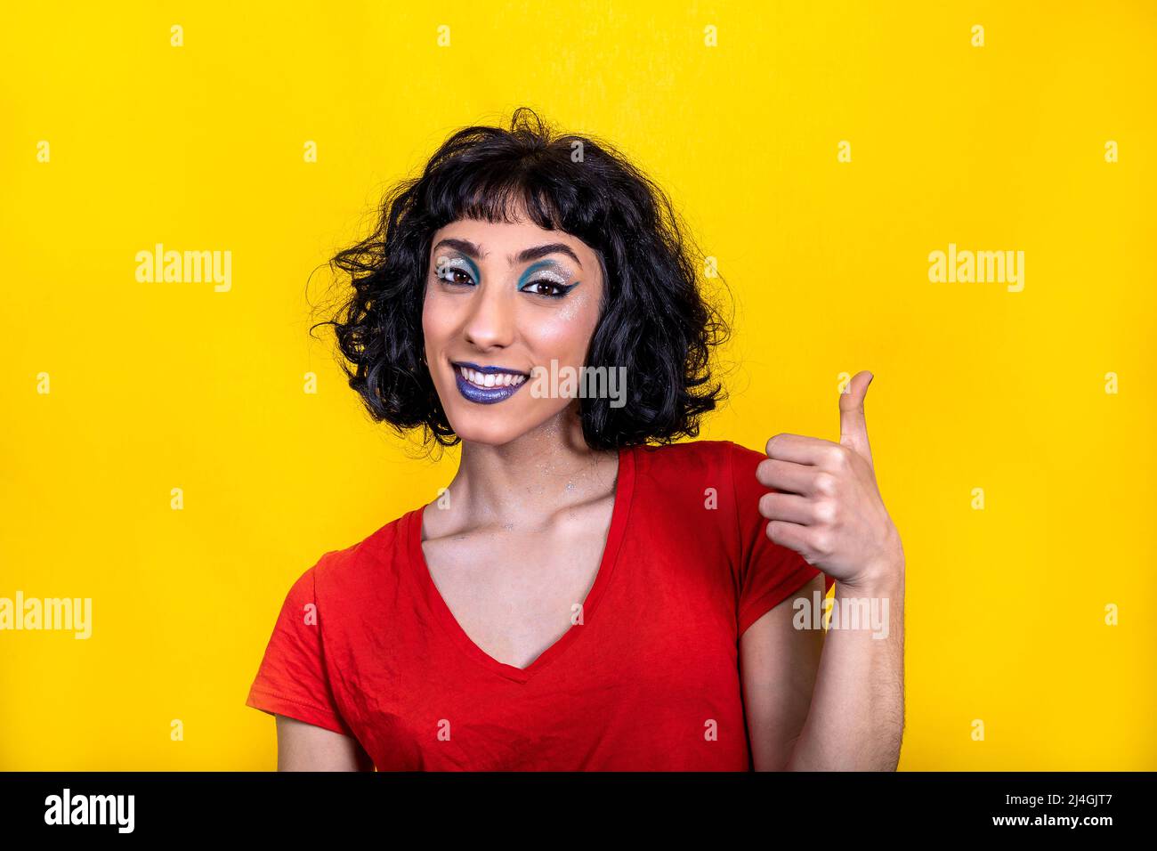 Smiling young woman in red t-shirt is showing thumb. Woman portrait with trendy look and bright colors on yellow background. Stock Photo