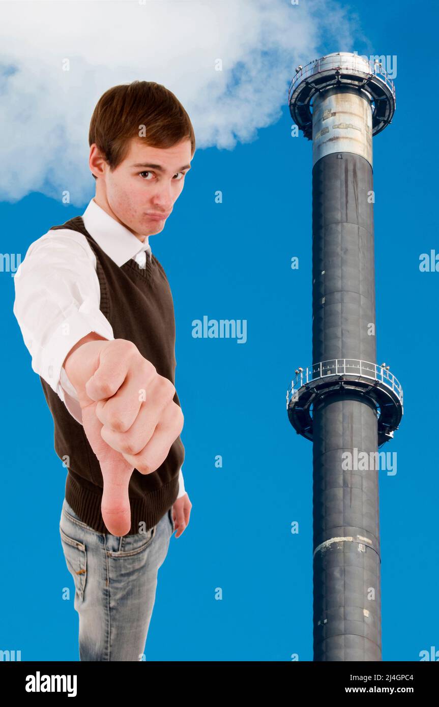Waist up view of a young white male showing thumbs down next to a smoking factory chimney against a blue sky. Stock Photo