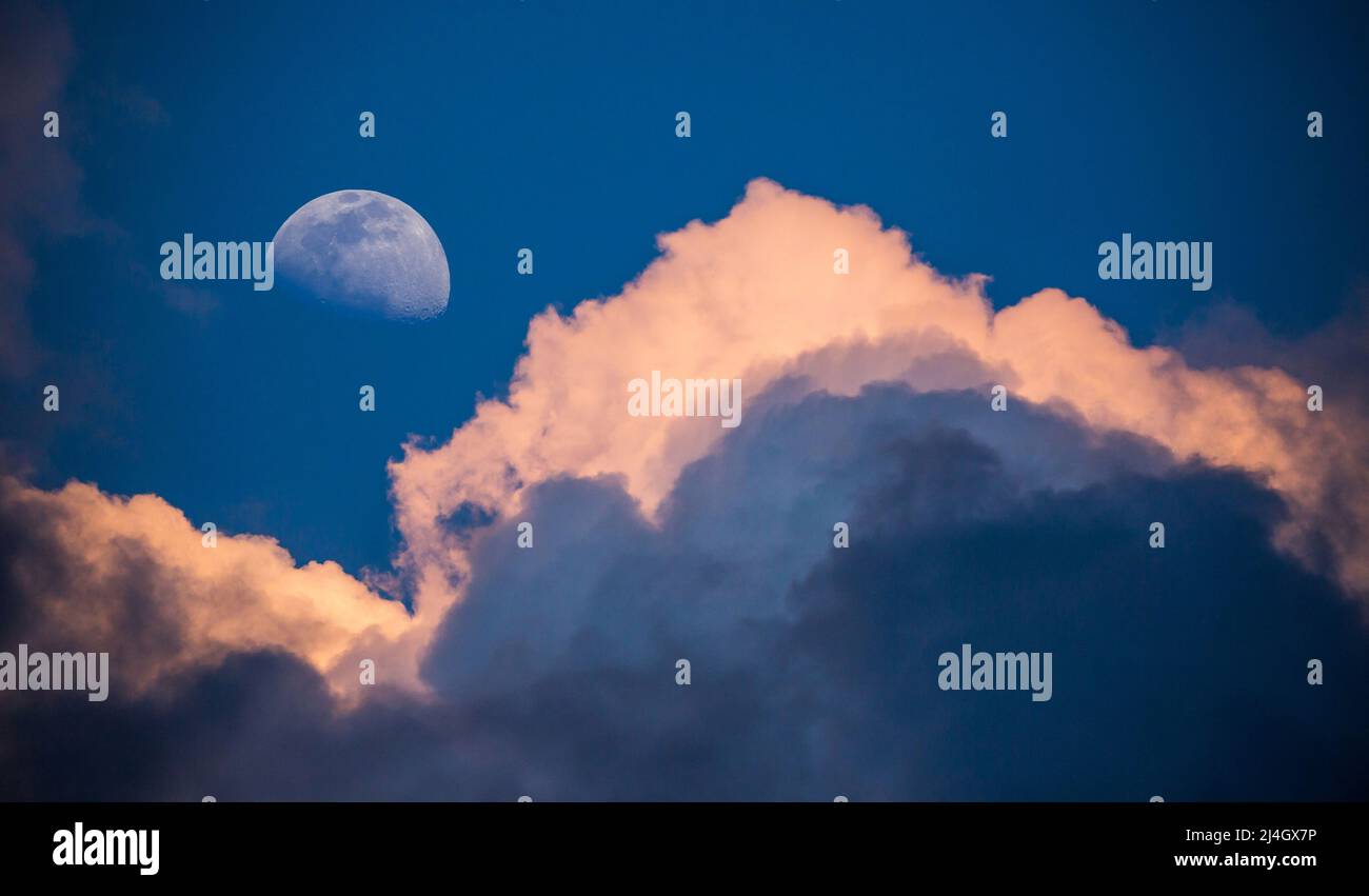 A waxing gibbous moon rises in a deep blue sky above gathering storm clouds lit by the setting sun. Stock Photo