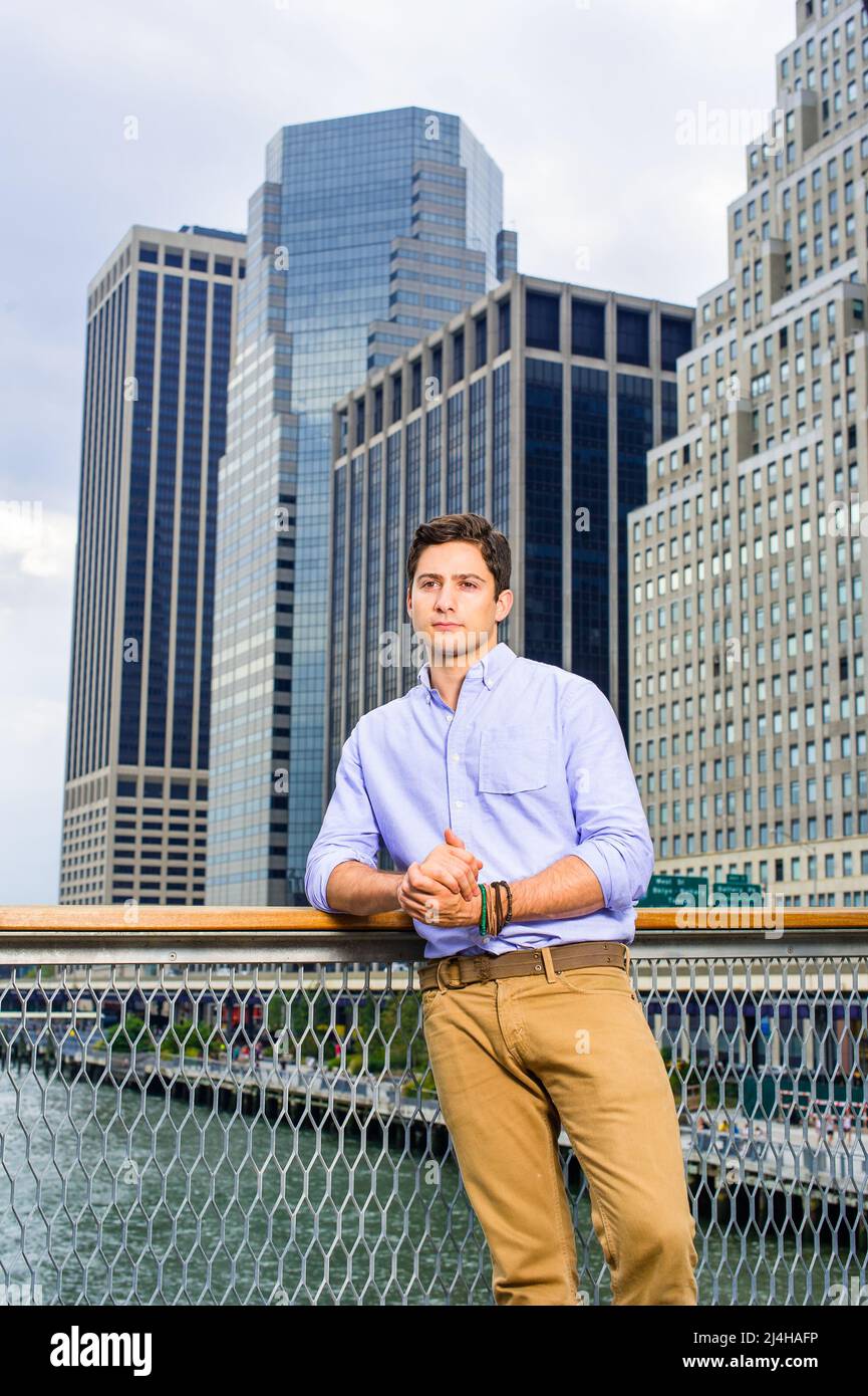 Dressing in a light blue shirt and dark yellow jeans, a young handsome guy is standing in the front of a business district, thinking. Stock Photo