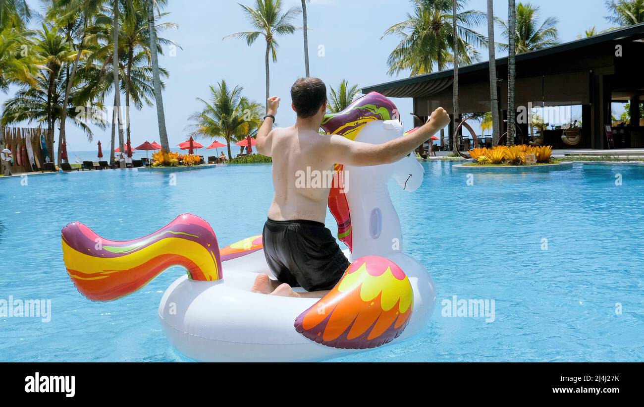 Funny man having fun, dancing on floating inflatable unicorn, tropical vacation Stock Photo