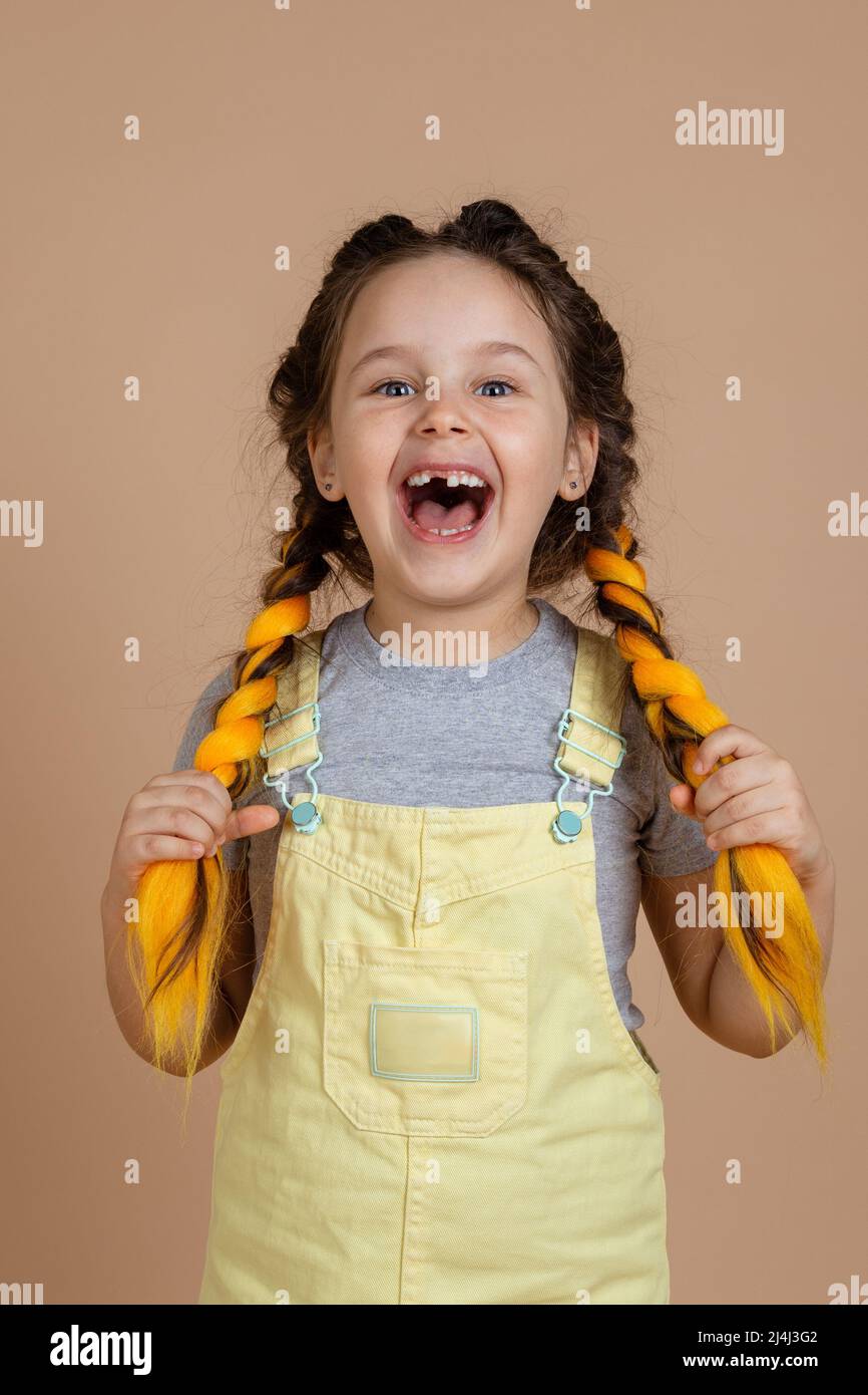 Fascinated small child touching yellow kanekalon pigtails with hands, smiling with opened mouth with missing tooth wearing yellow jumpsuit and gray t Stock Photo