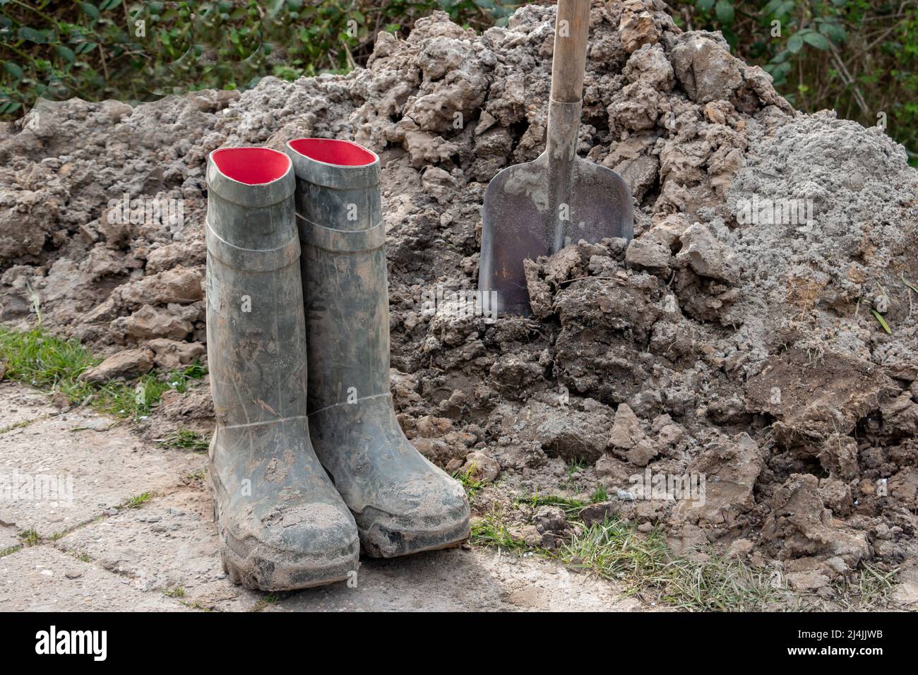 A shovel in a heap of clay soil, after hand-digging a piece of soil and having dirty green boots Stock Photo