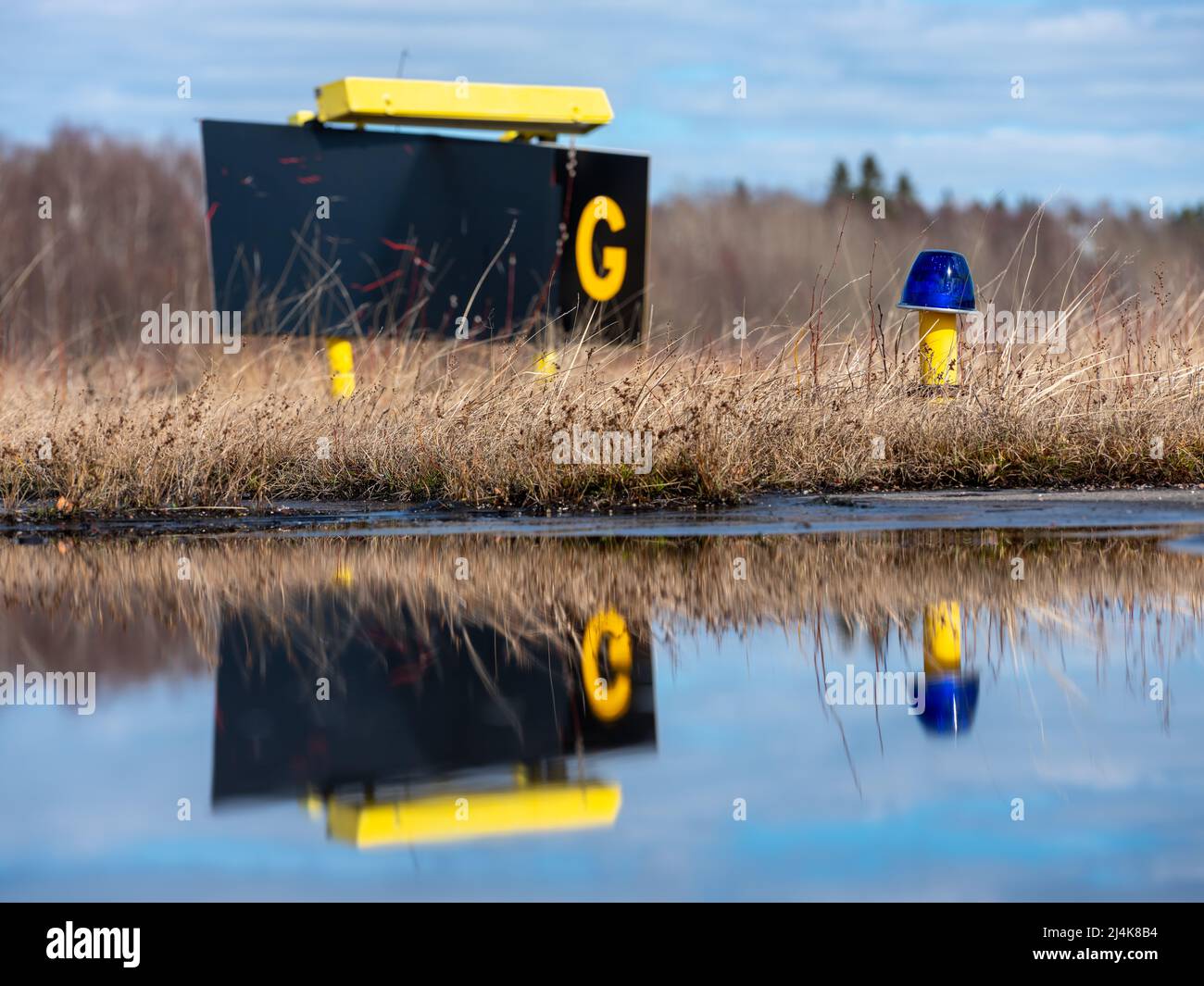 Helsinki / Finland - APRIL 16, 2022: Closeup of a blue airport taxiway light with taziway G sign in the background. Reflection on the water. Stock Photo