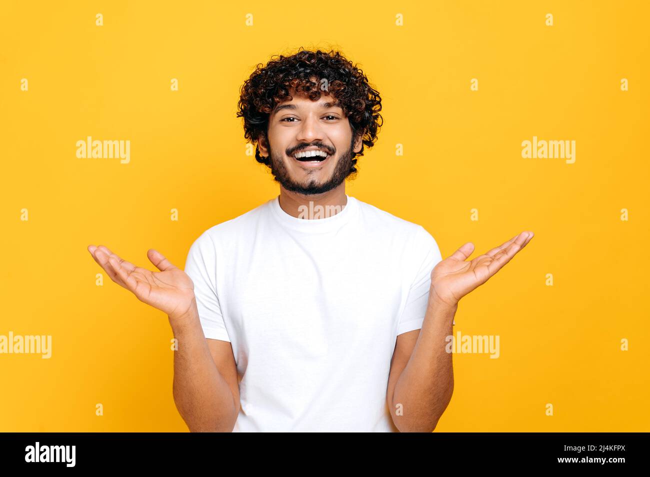 Joyful positive indian or arabian guy with curly hair, wearing white t-shirt, looks at camera happily, spreading his arms to the sides, standing over isolated orange background, looks at camera, smile Stock Photo