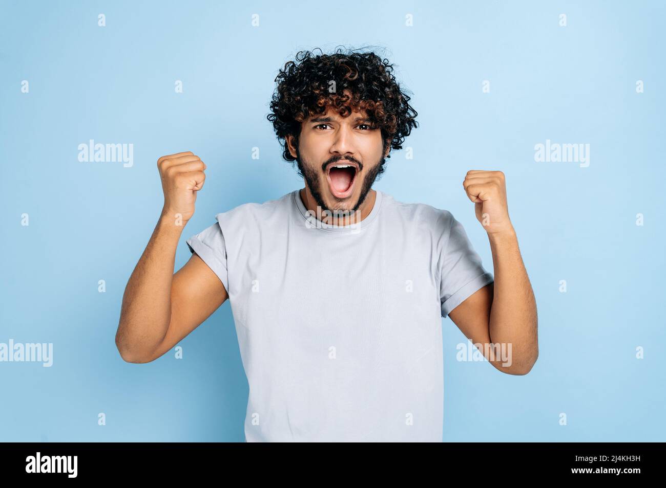 Win, triumph. Excited amazed indian or arabian guy, in t-shirt, rejoices in success, victory, win, gesturing with fists, looking at camera, smiling, standing on an isolated blue background, shouting Stock Photo