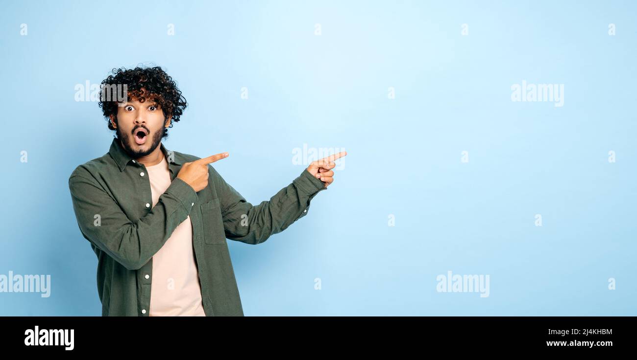 Panoramic photo of amazed shocked indian or arabian guy in casual wear, points fingers to the side at mock-up empty space, stands on isolated blue background, looks surprised at the camera, astonished Stock Photo