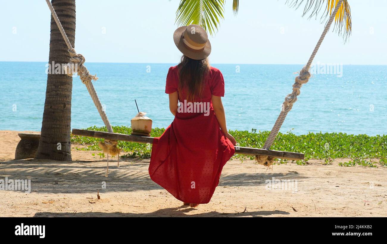 Travel woman sit, rest on swing at exotic beach, drink fresh coconut