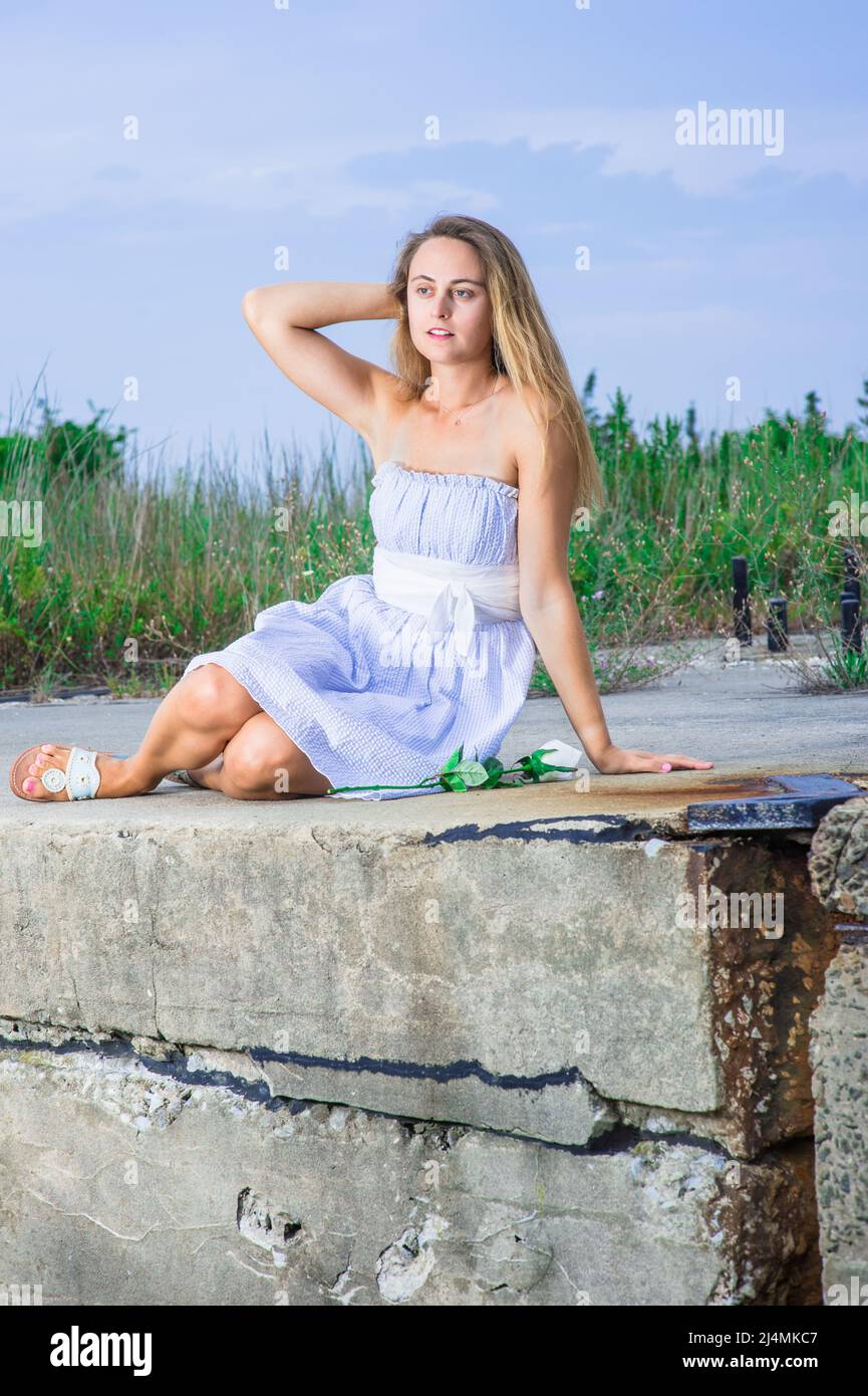 A pretty blonde girl, dressing in a white blue dress, white waistband and  sandals, is sitting on a high land, thoughtfully looking forward. A white r  Stock Photo - Alamy