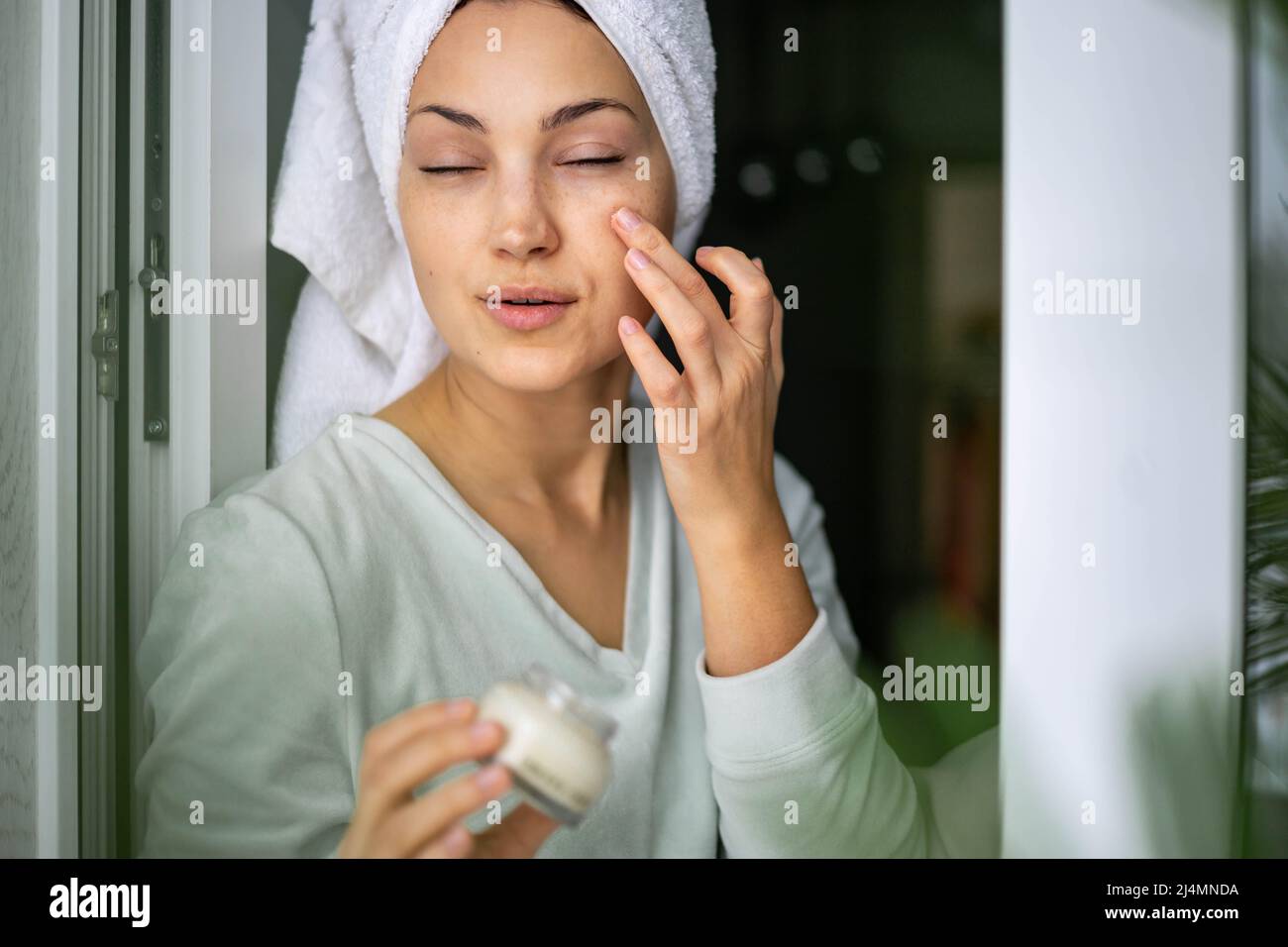 Happy domestic woman applying anti aging cream from jar on face looking at mirror Stock Photo
