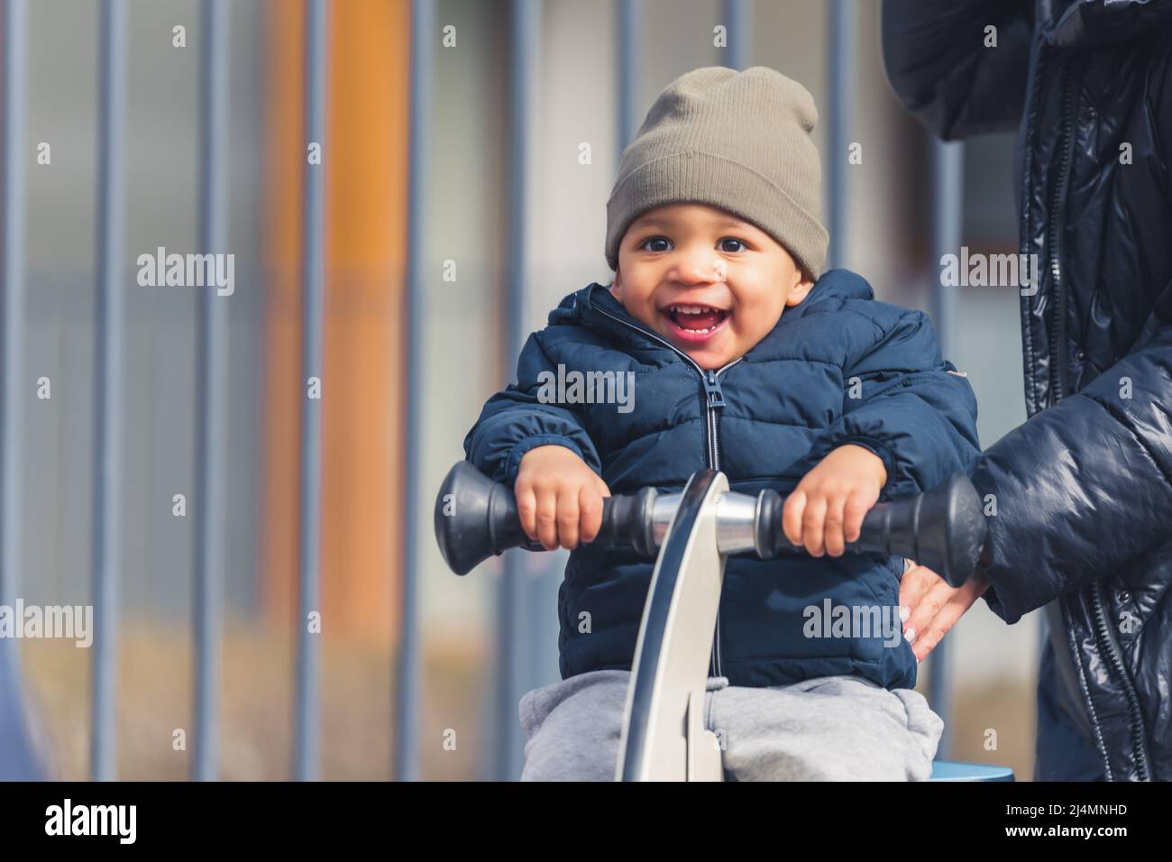Outdoor portrait of an extremely happy smiley young biracial toddler in a gray hat and warm coat sitting on a playground spring rider and having fun. High quality photo Stock Photo