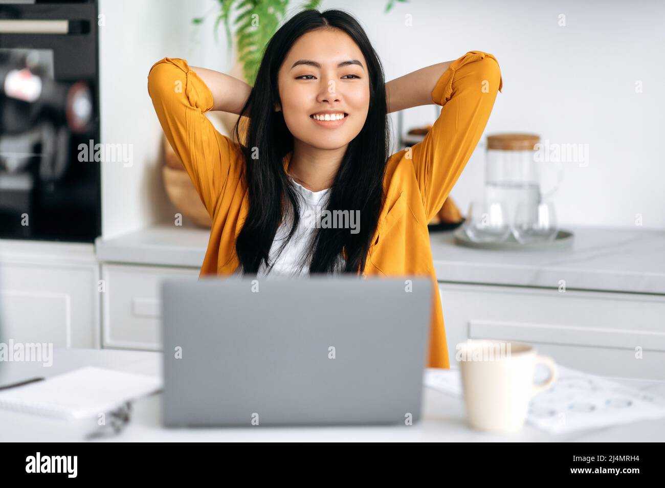 Happy Chinese girl, freelancer, working from home, sits in the kitchen at the workplace, takes a break from work, puts her hands behind her head, looks to the side, dreams of rest, thinks, smiles Stock Photo