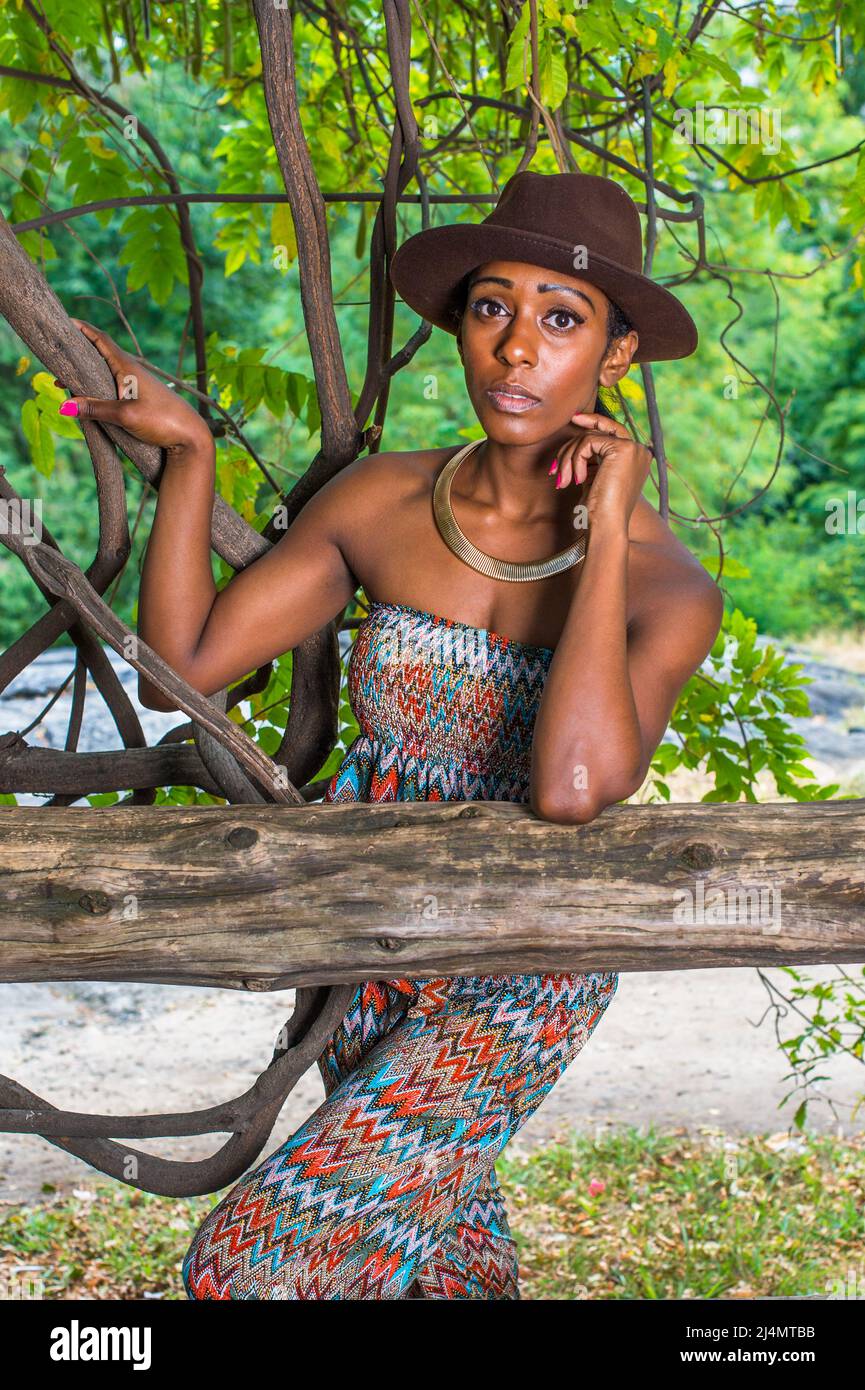 Wearing a brown outback hat,a golden neck ring, a strapless fashionable, contemporary style dress top, pants , a pretty black woman is standing outsid Stock Photo