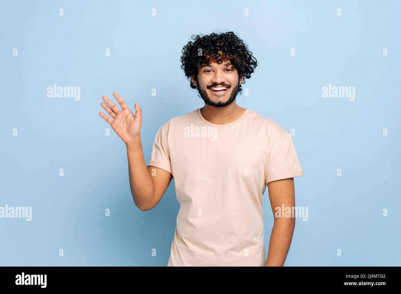 Handsome positive arabian or indian curly-haired guy, wearing casual t-shirt, standing on isolated blue background, looking at the camera, waving hand, welcome sign, smiling friendly Stock Photo