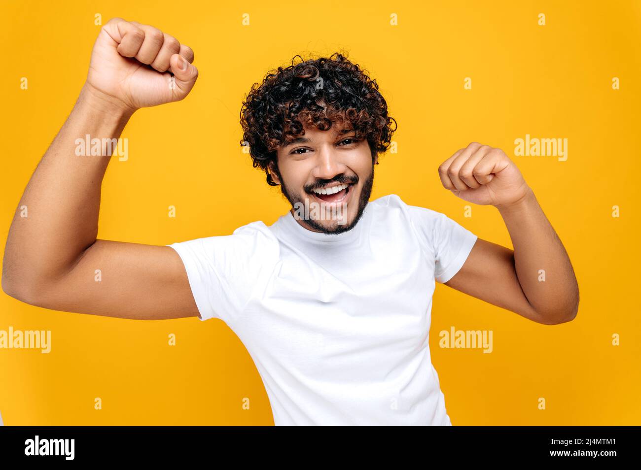 Joyful positive indian or arabian guy with curly hair, wearing white t-shirt, looks at camera happily, spreading his arms to the sides, standing over isolated orange background, looks at camera, smile Stock Photo