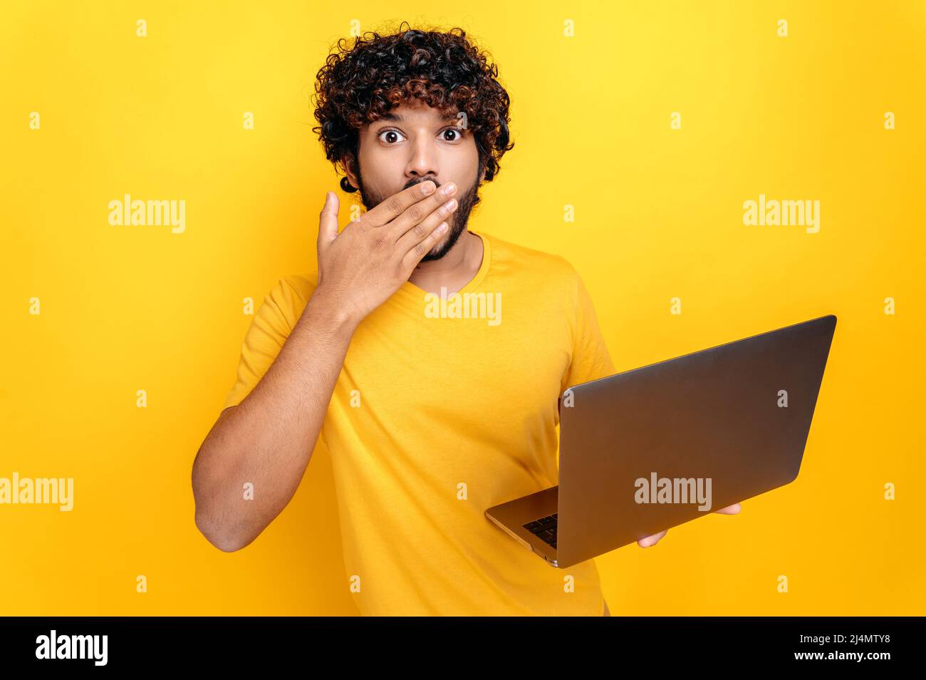 Shocked indian or arabian curly-haired guy, holding an open laptop in his hand, looks surprised at the camera, covering mouth with hand, while standing on isolated orange background Stock Photo