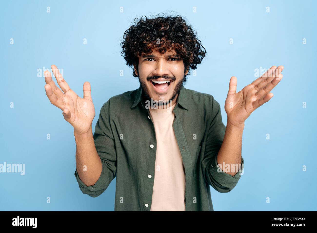 Discouraged Indian or Arabian confused curly-haired young man, emotionally gesturing with his hands, looking inquiringly at the camera, standing on an isolated blue background Stock Photo