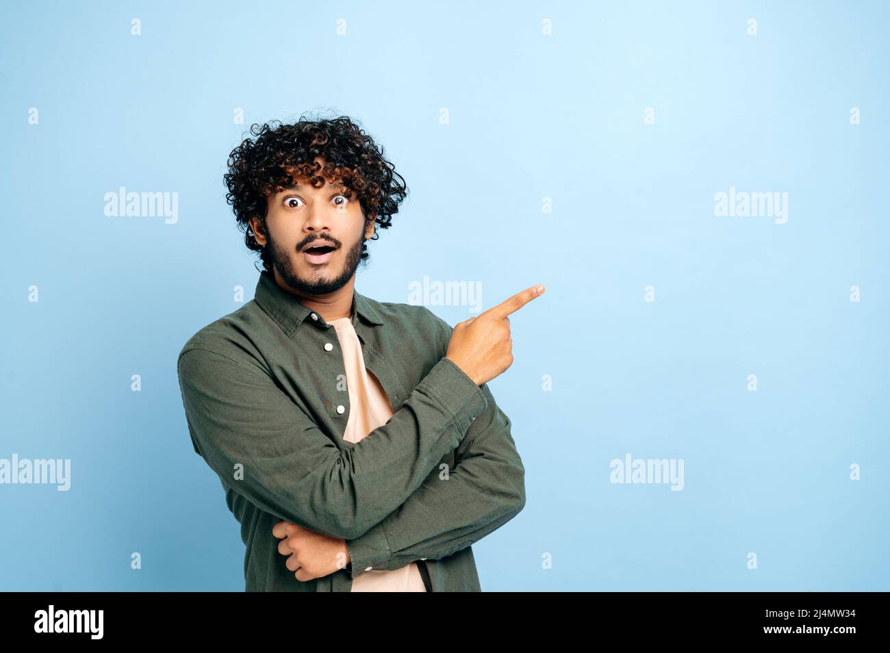 Surprised shocked indian or arabian guy in casual clothes, amazed looks at camera and points finger to the side at space for presentation, stands on isolated blue background, smile. Copy space concept Stock Photo