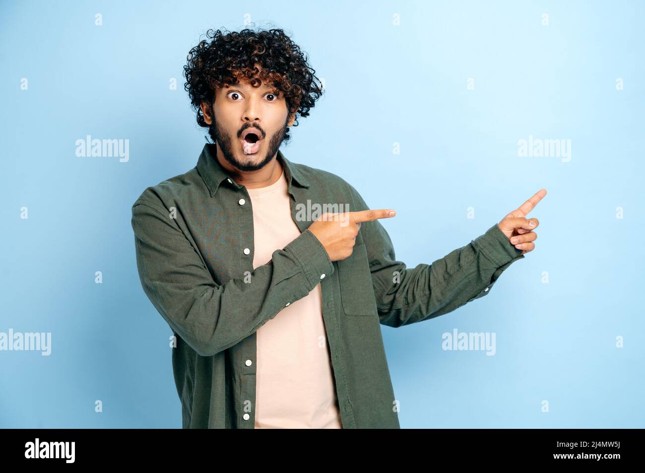 Amazed shocked indian or arabian curly haired guy in casual wear, points fingers to the side at mock-up empty space, stands on isolated blue color background, looks surprised at the camera, astonished Stock Photo