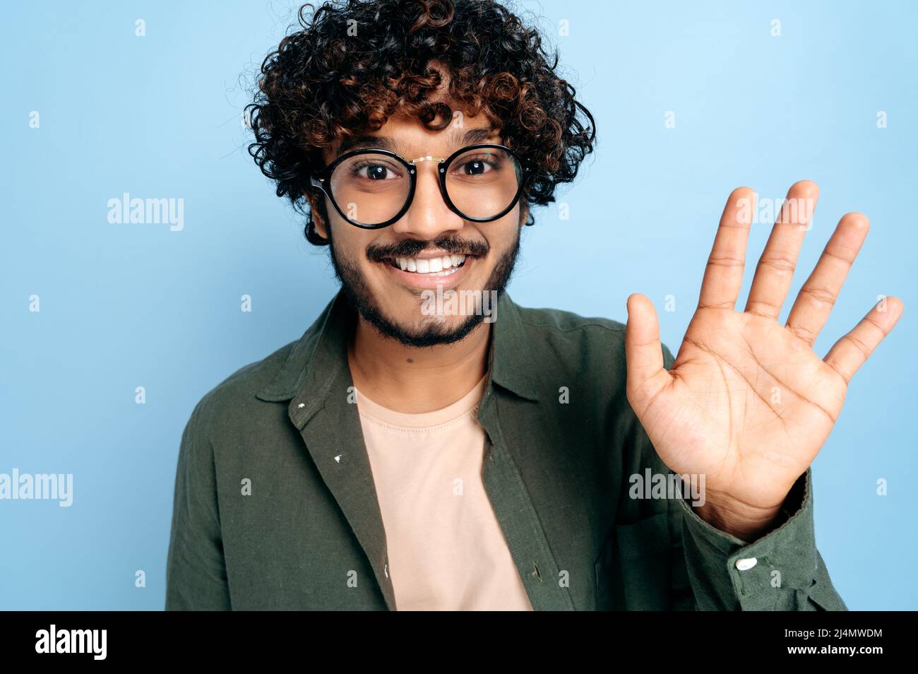 Close-up of charismatic attractive positive curly indian or arabian guy in casual shirt with glasses, looking at camera, smiling happily, waving, greeting gesture, stand on an isolated blue background Stock Photo