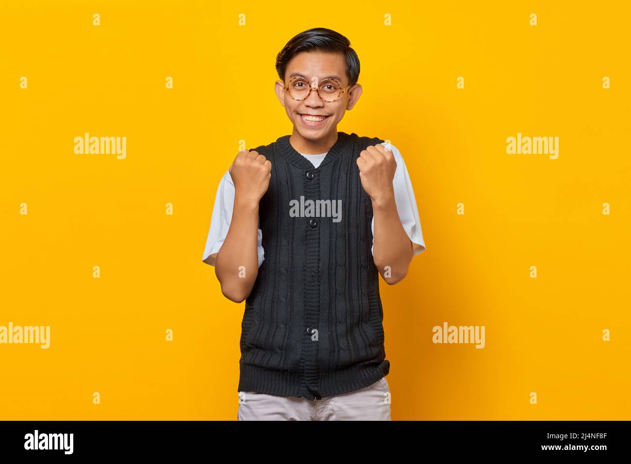 Portrait of excited young Asian man celebrating success with raised arms on yellow background Stock Photo