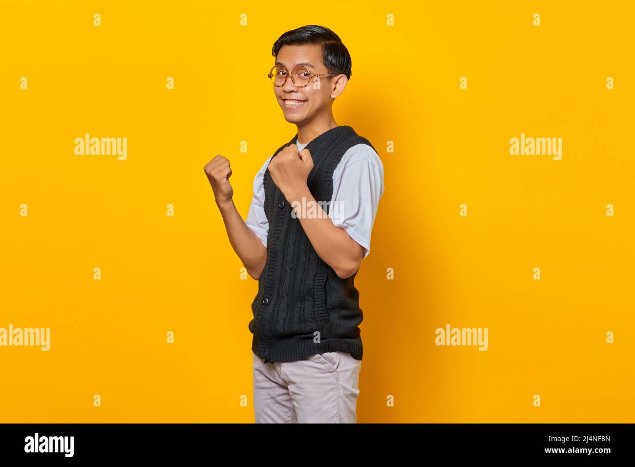 Portrait of excited young Asian man celebrating success with raised arms on yellow background Stock Photo