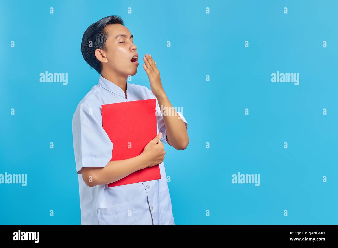 Young Asian male nurse holding folder feeling sleepy and covering mouth with hand on blue background Stock Photo